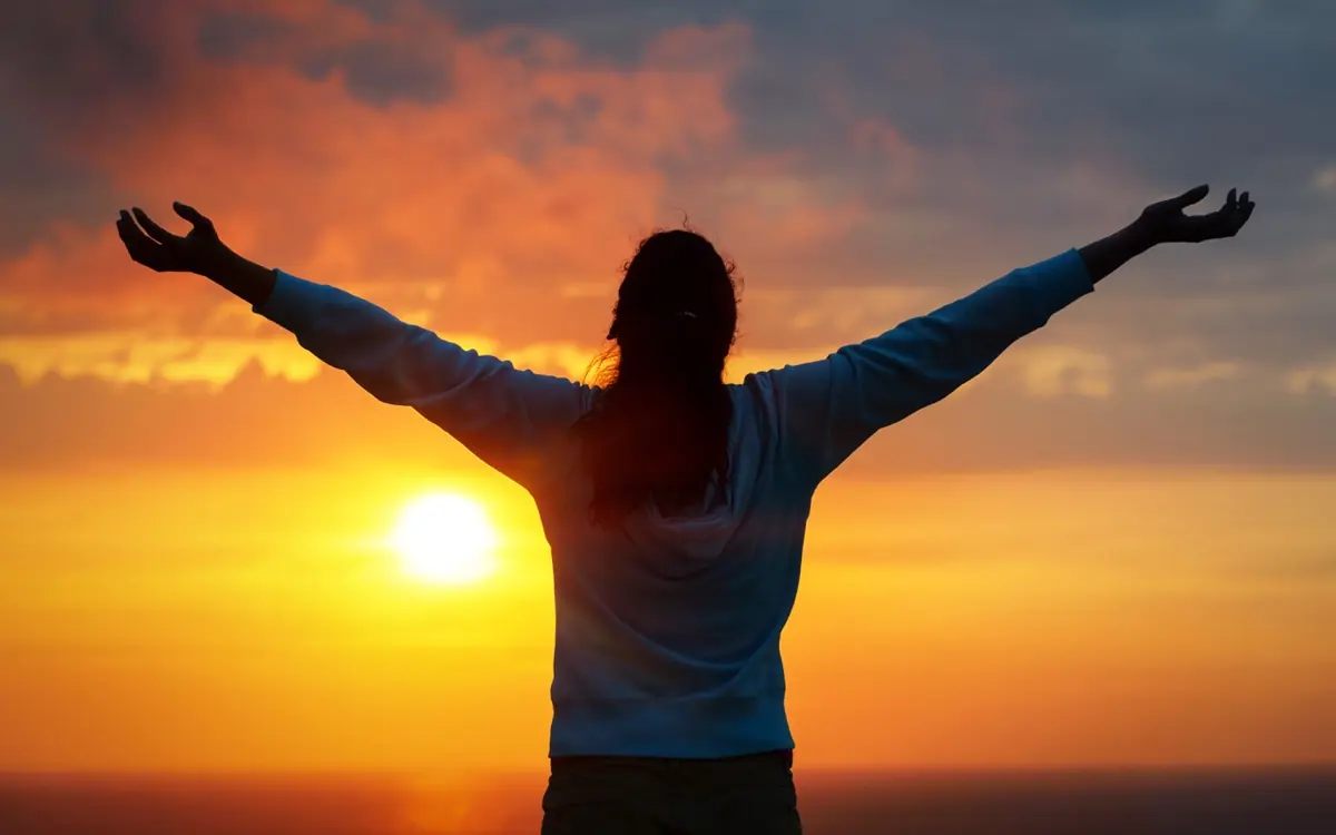 A person opens their arms toward the evening light outdoors.