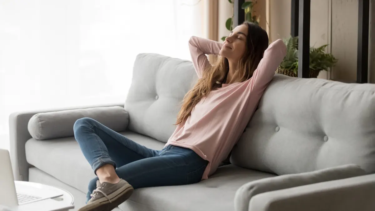 A woman stretches while sitting on a sofa in a bright room.