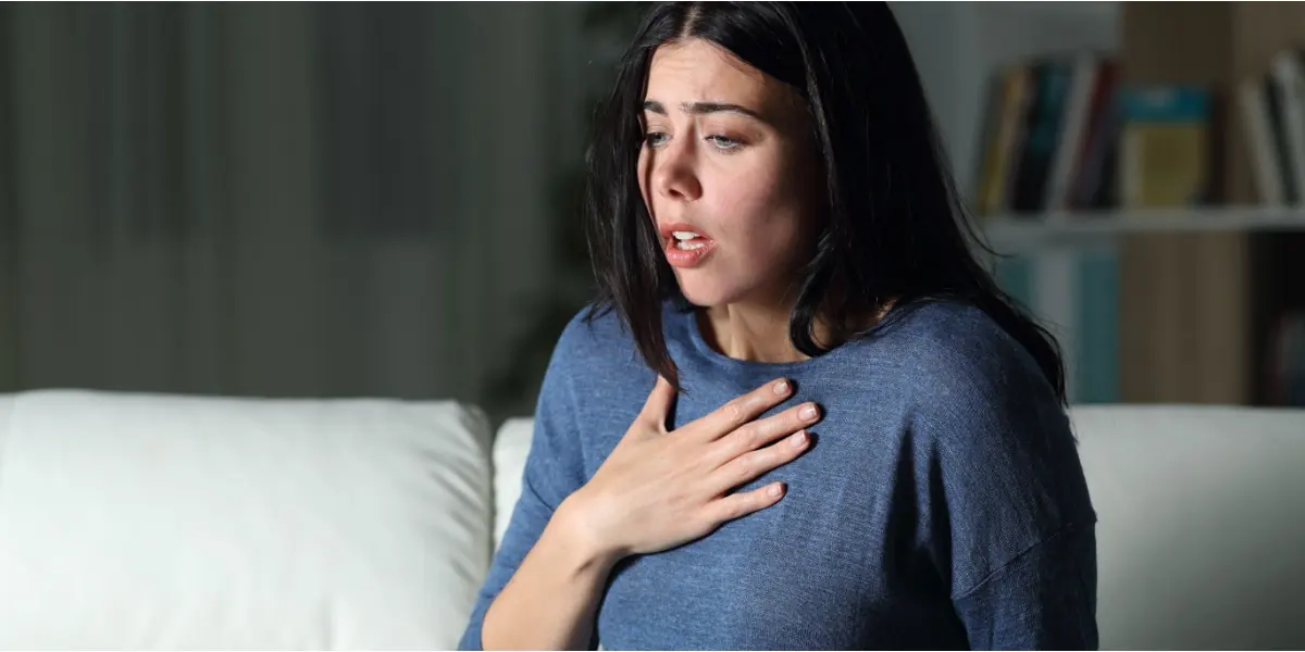 A woman rests a hand on her chest while sitting indoors.