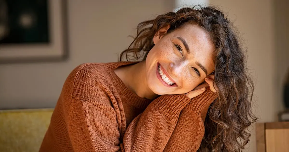 A smiling woman rests her head on her shoulder in a bright room.