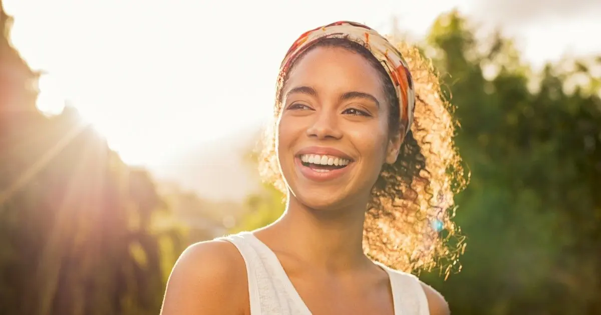 A smiling woman enjoys warm sunlight outdoors.