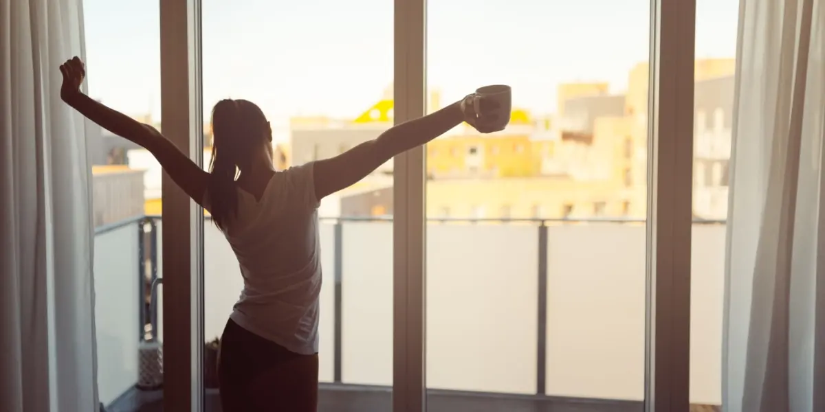 A person stretches their arms near an open window in morning light.