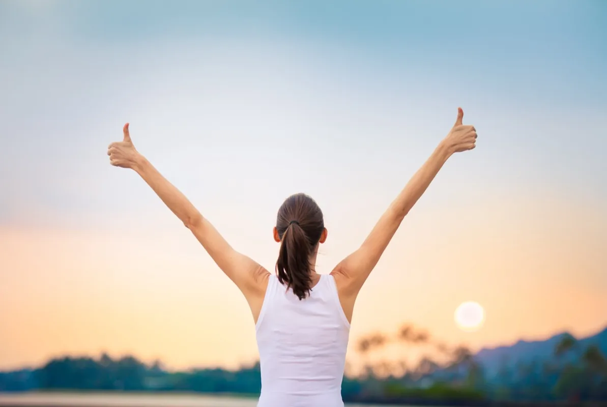 A woman lifts her arms while looking toward a warm evening sky.