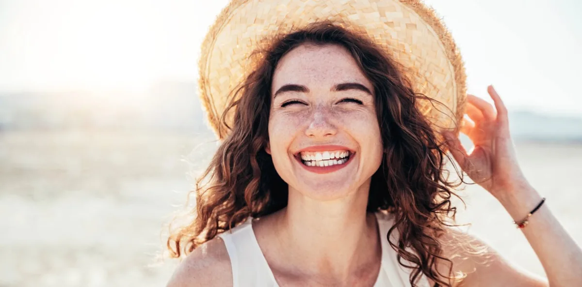 A woman smiles in the sunshine while wearing a straw hat.