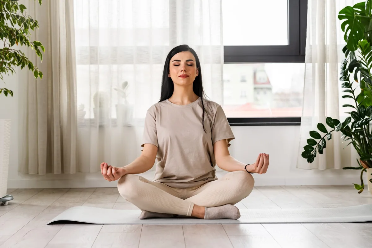 A woman sits cross-legged with her eyes closed in a bright room.