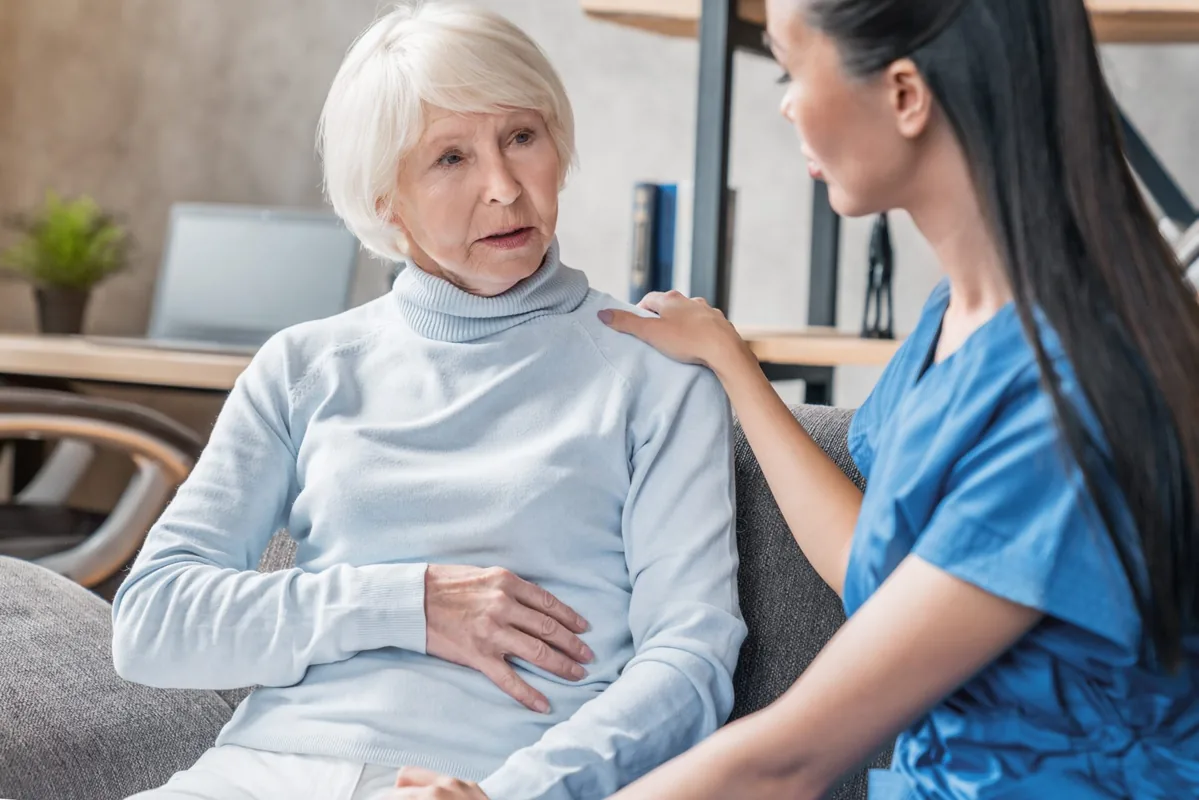 An older patient speaks with a healthcare professional beside a window.