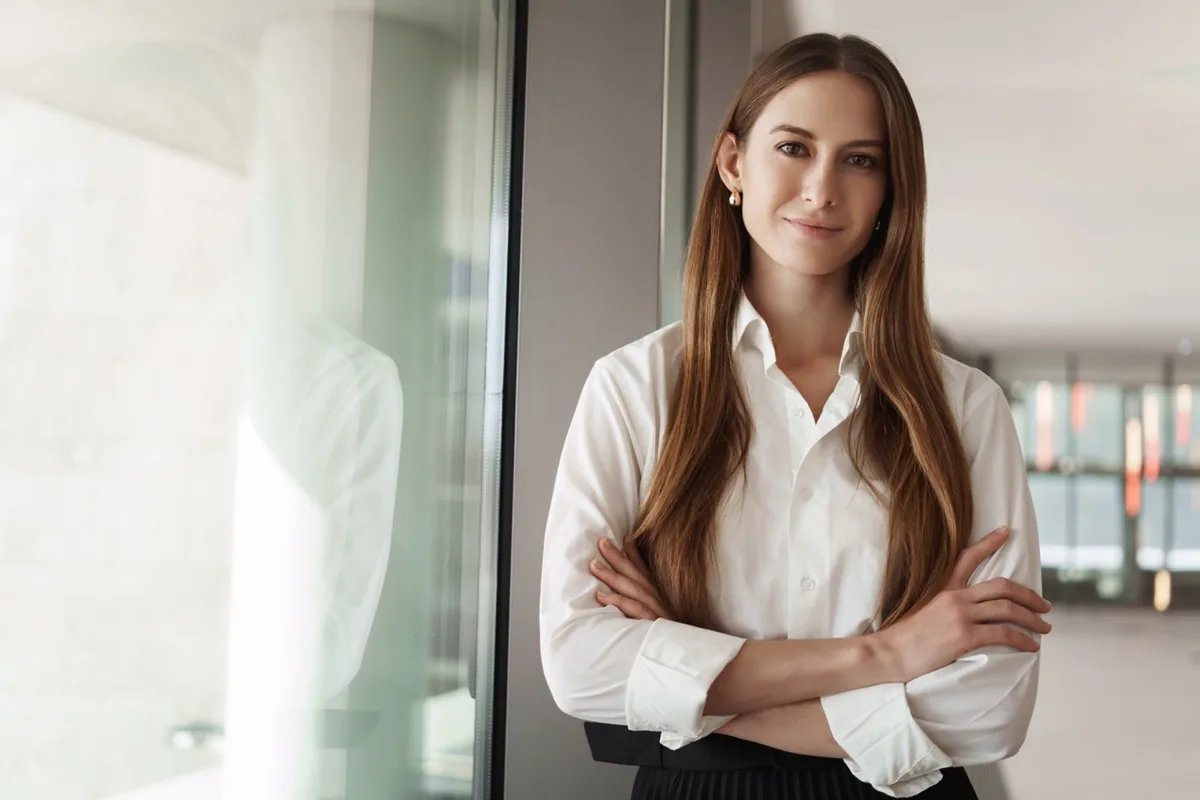 A woman stands confidently in a bright room with her arms folded.