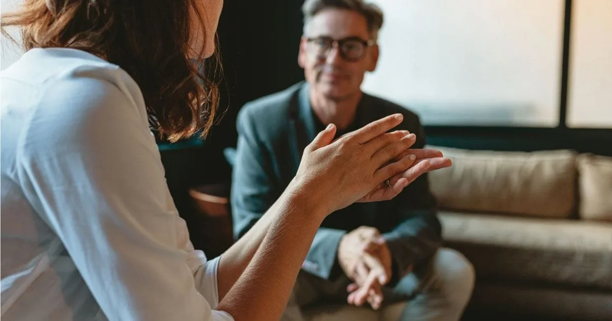 A man speaks with a woman during a quiet conversation indoors.