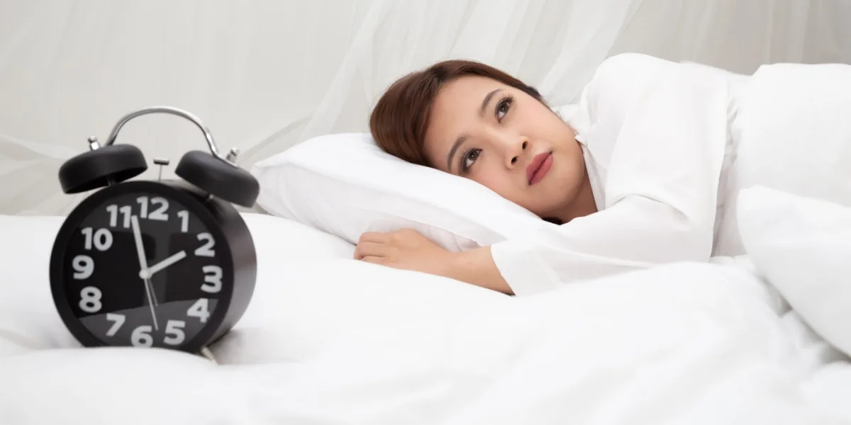 A woman lies awake in bed beside an alarm clock.