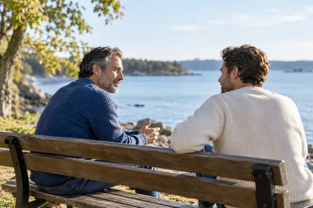 Two men talking on a park bench overlooking water.