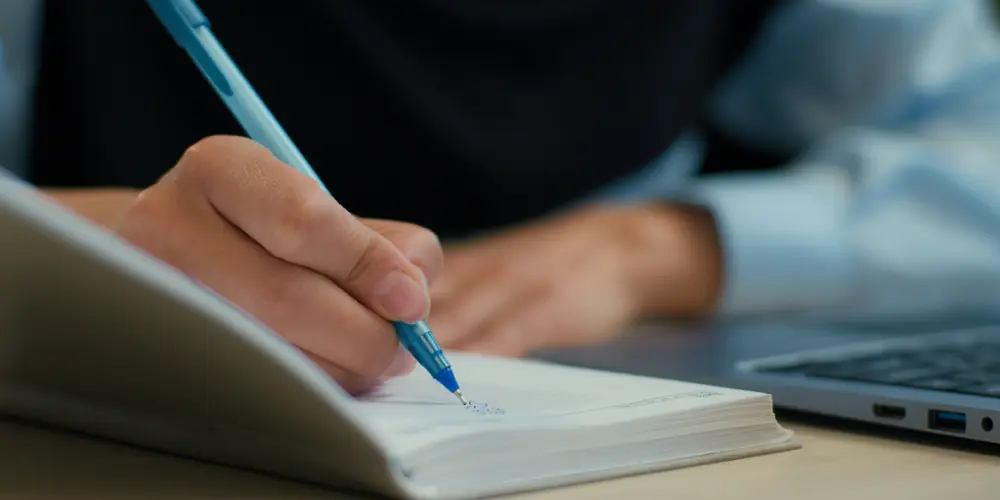 A person writes in a notebook beside an open laptop.