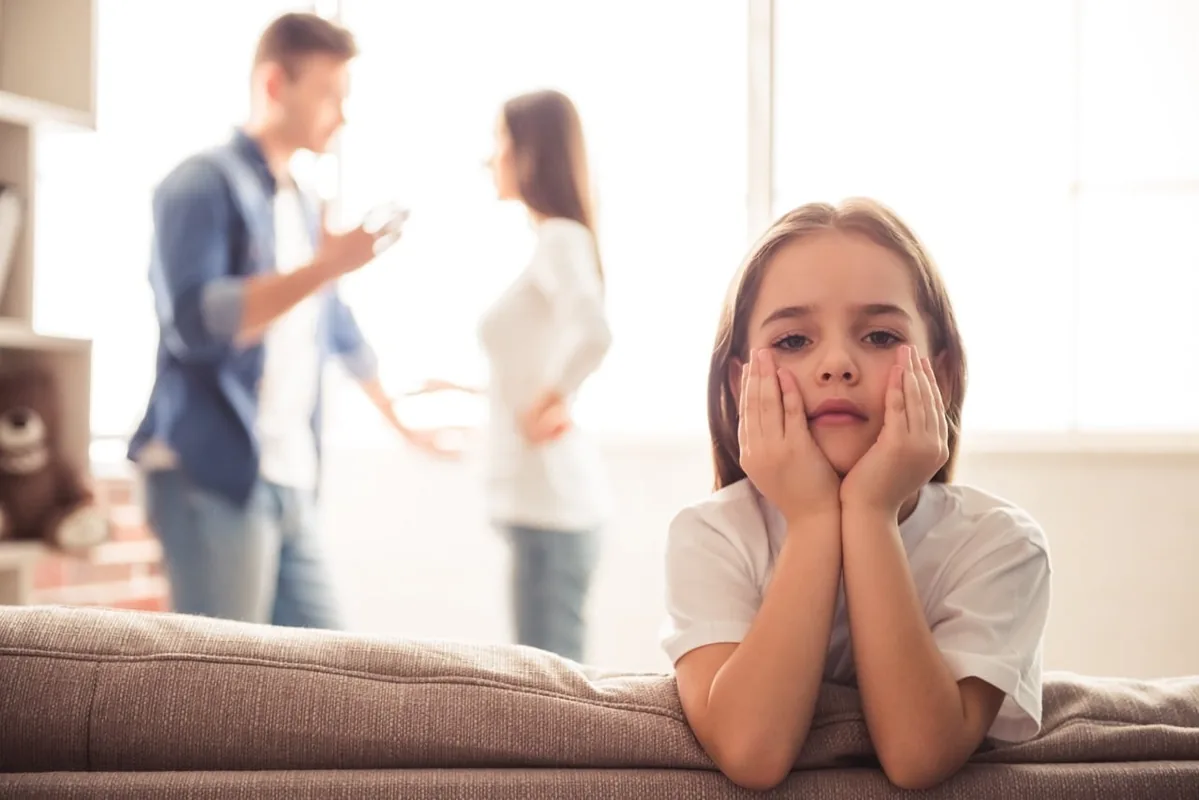 A child rests her chin in her hands while adults speak in the background.