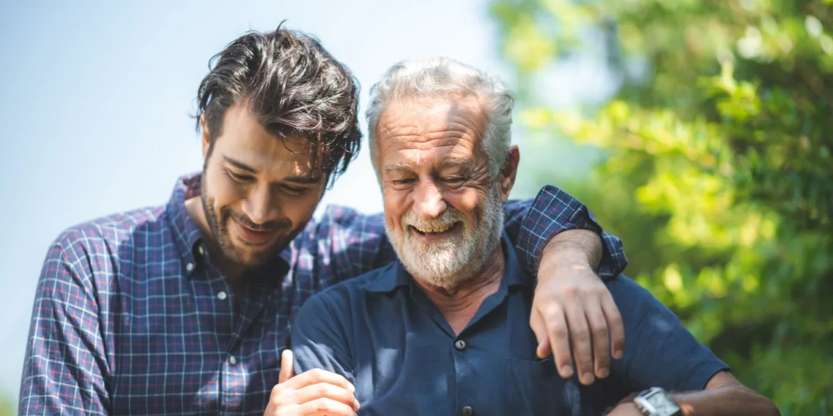 An older man rests his arm around a younger man outdoors.