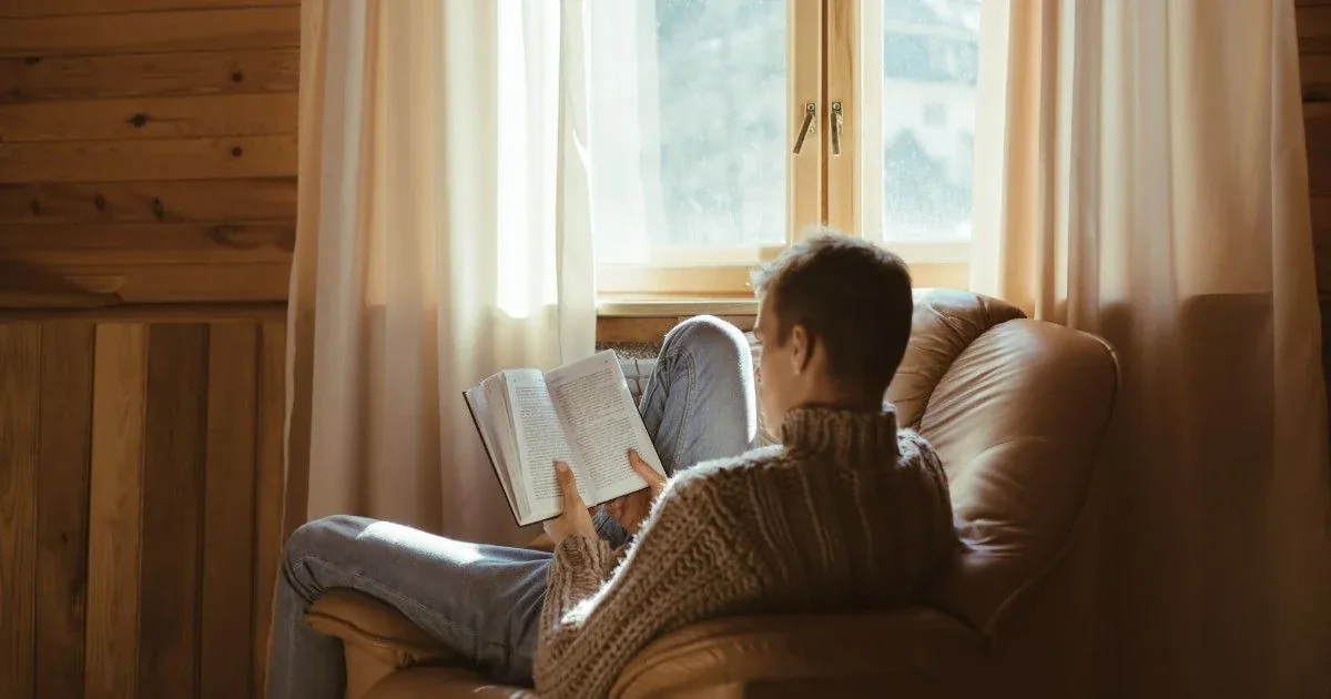 A person reads quietly on a sofa beside a window.