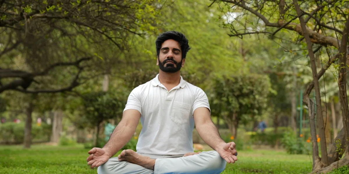 A man sits cross-legged beneath leafy trees with his eyes closed.