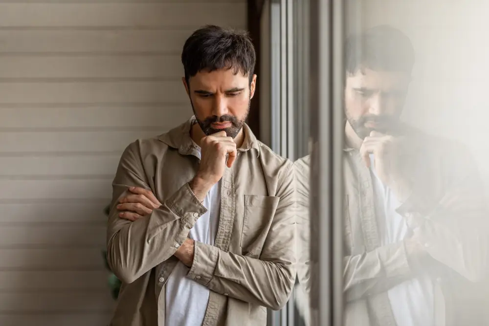 A man sits with clasped hands beside a window.