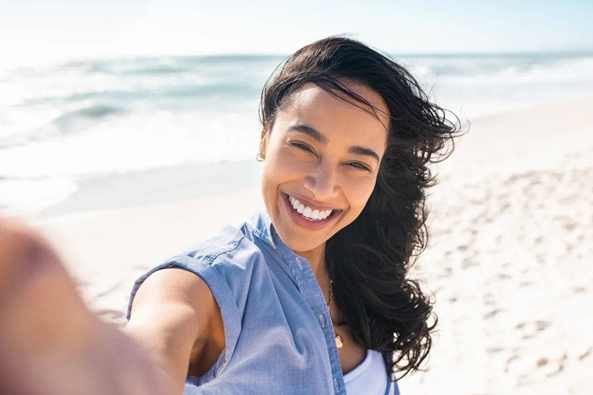A woman smiles in the sunlight beside the sea.