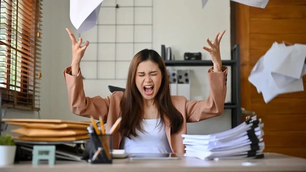 A woman at a desk reacts to an overwhelming pile of paperwork.