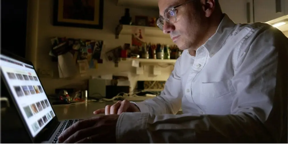A man sits at a laptop in a dim room.