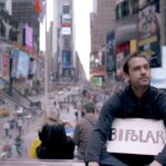 Ross McKenzie holds a sign bearing 'Bipolar' while filming 'Bipolarized' in New York City, symbolizing his commitment to challenging the modern mental health care model