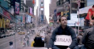 Ross McKenzie holds a sign bearing 'Bipolar' while filming 'Bipolarized' in New York City, symbolizing his commitment to challenging the modern mental health care model