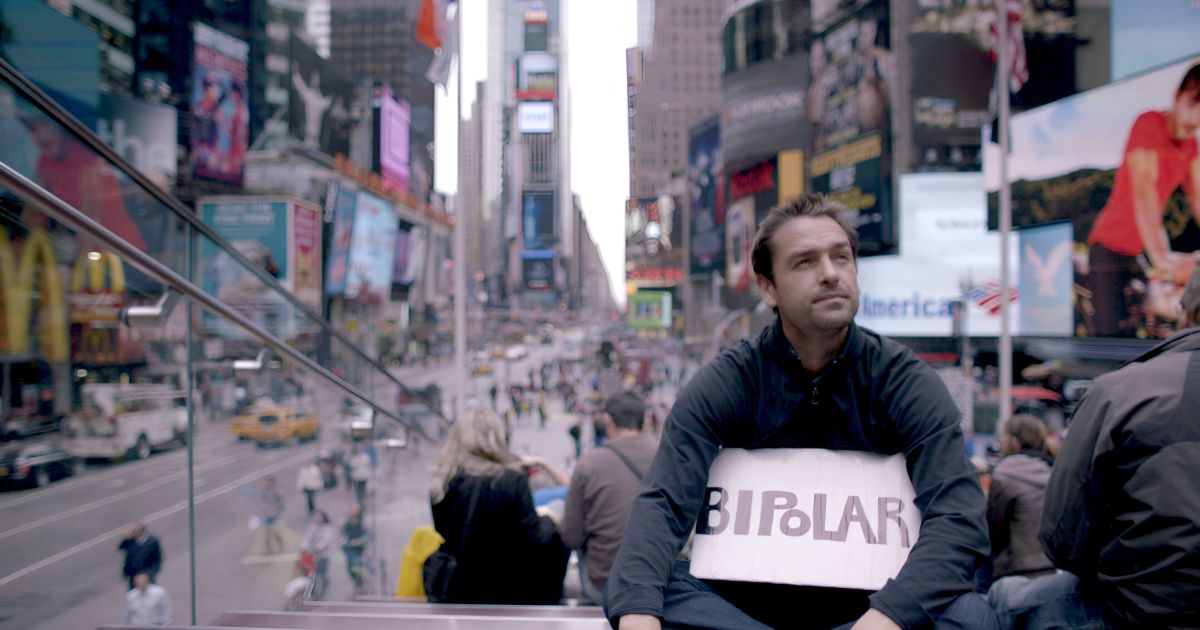 Ross McKenzie holds a sign bearing 'Bipolar' while filming 'Bipolarized' in New York City, symbolizing his commitment to challenging the modern mental health care model